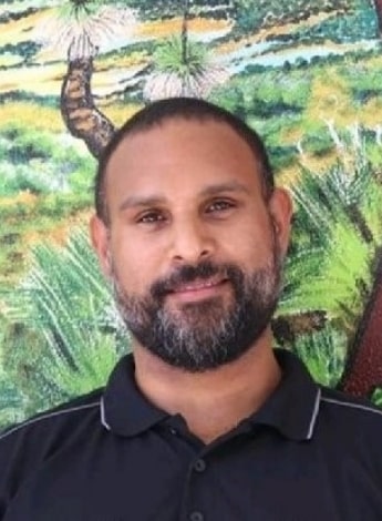A man with a closely trimmed beard is wearing a black polo shirt and standing in front of a colourful background