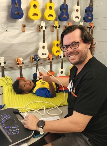 A man wearing a black t shirt holds an echocardiogram probe to the chest of a young Aboriginal boy. The boy is lying on a bed in front of a wall full of ukeleles. 