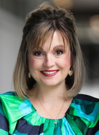 "A woman wearing a green and blue dress is smiling at the camera. She has shoulder length straight blonde hair." 