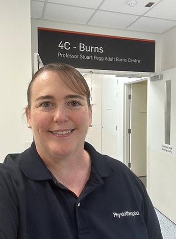 "A woman with her brown hair pulled back is standing in a corridor. The tag on her navy top reads physiotherapist. There is a sign above her that reads Burns."
