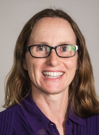 A woman with shoulder length brown hair and dark-rimmed glasses is smiling at the camera. 