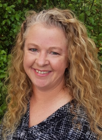 A woman with long curly blonde hair is standing in front of greenery