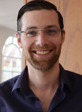 A man with short brown hair and a dark shirt is looking at the camera