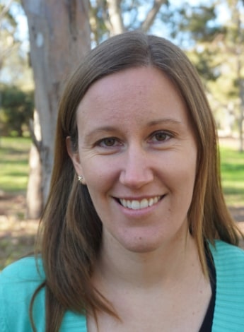 "A woman with long brown hair is smiling at the camera. She is wearing a blue top and is standing outside in front of a tree."