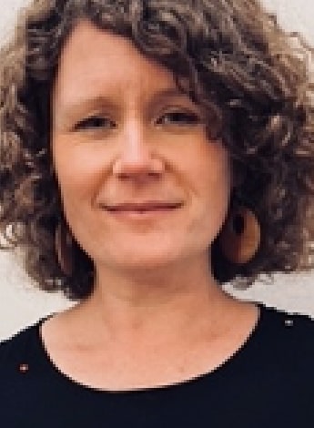 A woman with curly brunette hair is wearing a black top and looking at the camera. 