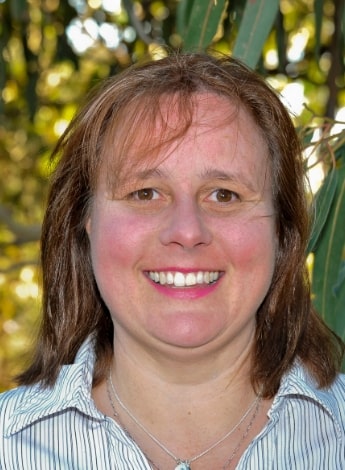 ""A woman with shoulder length brown hair is smiling at the camera. She is wearing a striped white shirt and is standing outside in front of some plants.""