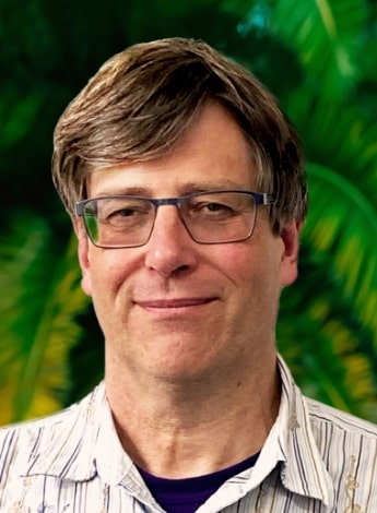 "A man wearing square-framed glasses and with brown hair is standing in front of plants and looking at the camera. "