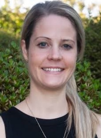 "A woman with blonde hair over one shoulder is smiling at the camera. She is wearing a black top and is standing outside in front of some plants"