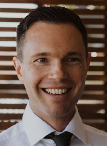 "A man wearing a white shirt and a black tie is standing in front of wooden window shutters."