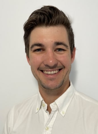 "A man with dark hair is smiling at the camera. He is wearing a white shirt."