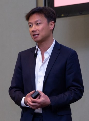 "An Asian man wearing a blue suit and a white shirt, is standing up holding a small device"