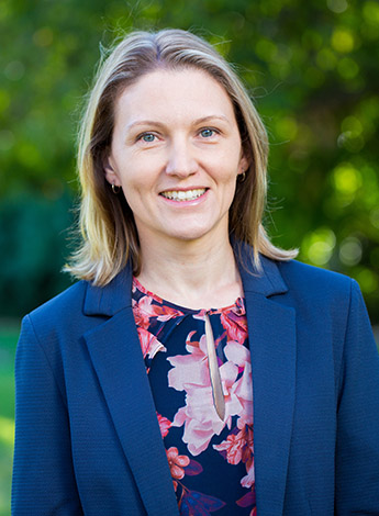 A blonde woman wearing a floral shirt and a blue blazer looks at the camera. She is standing outside in front of a green background. 