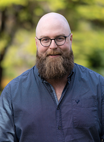 A bald bearded man wearing a blue shirt  is standing outside in front of greenery smiling at the camera. 