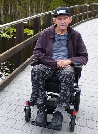 An older man is wearing a hat and sitting in a wheelchair. He is outside. 