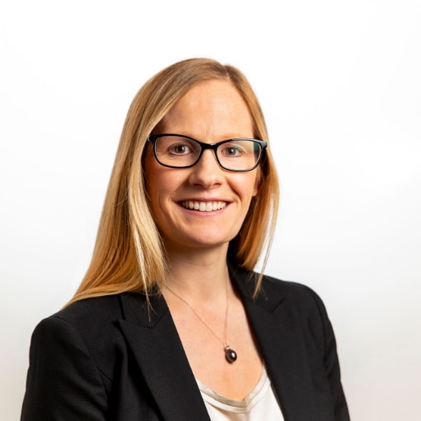 "A woman with long blonde hair is smiling at the camera. She is wearing a black blazer and black framed glasses."