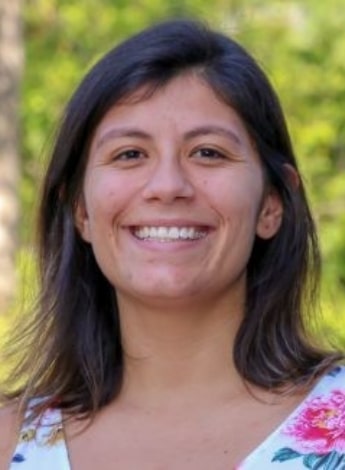 "A woman with shoulder length brown hair is standing in front of greenery and smiling at the camera "