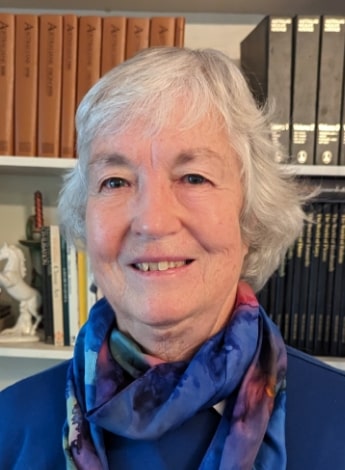 "A woman with short grey hair with a blue scarf around her neck is in front of bookshelves and smiling at the camera"