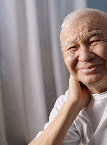 "An older man wearing a white t shirt is holding his neck"